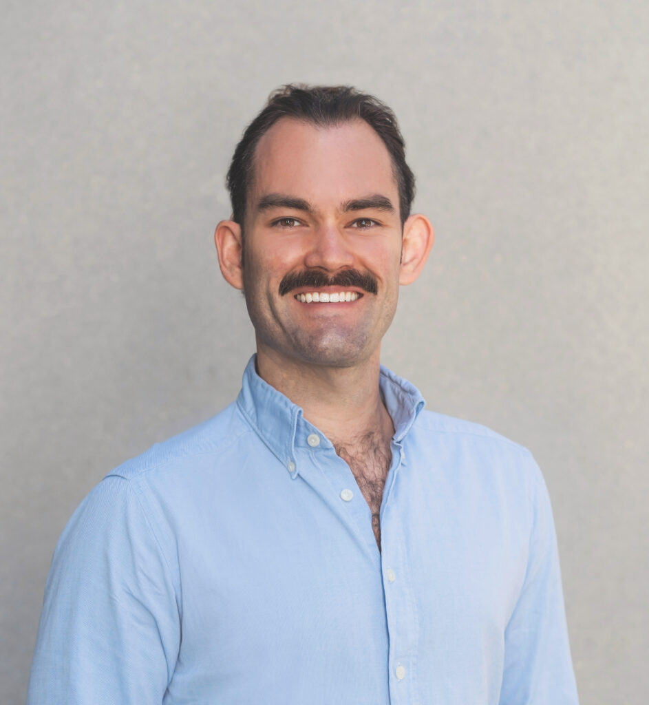 A man in a blue button up shirt with a moustache and his hair tied back