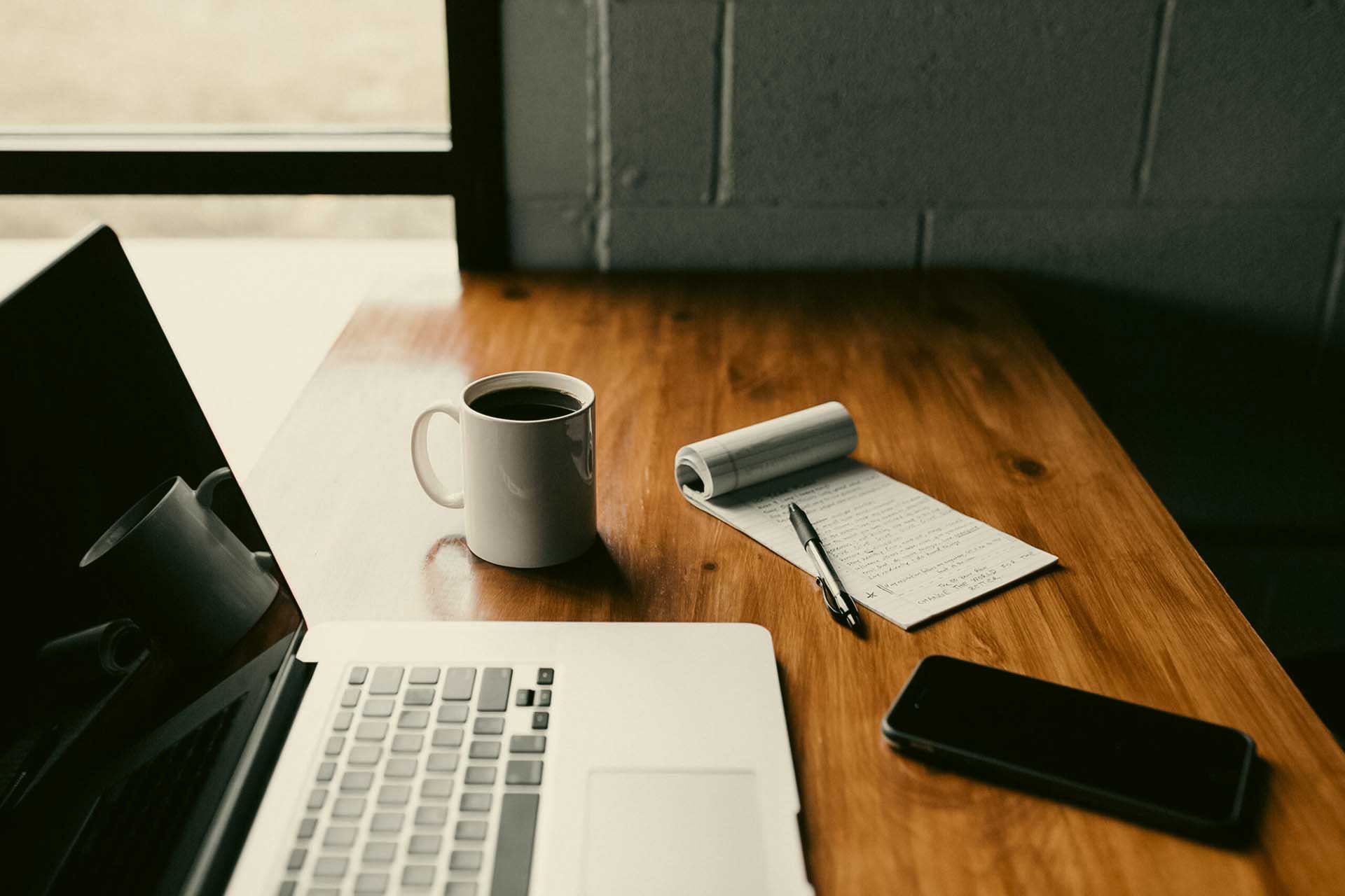 A silver Mac laptop sitting on a brown wooden table alongside a white coffee mug, notepad, pen, and cellphone