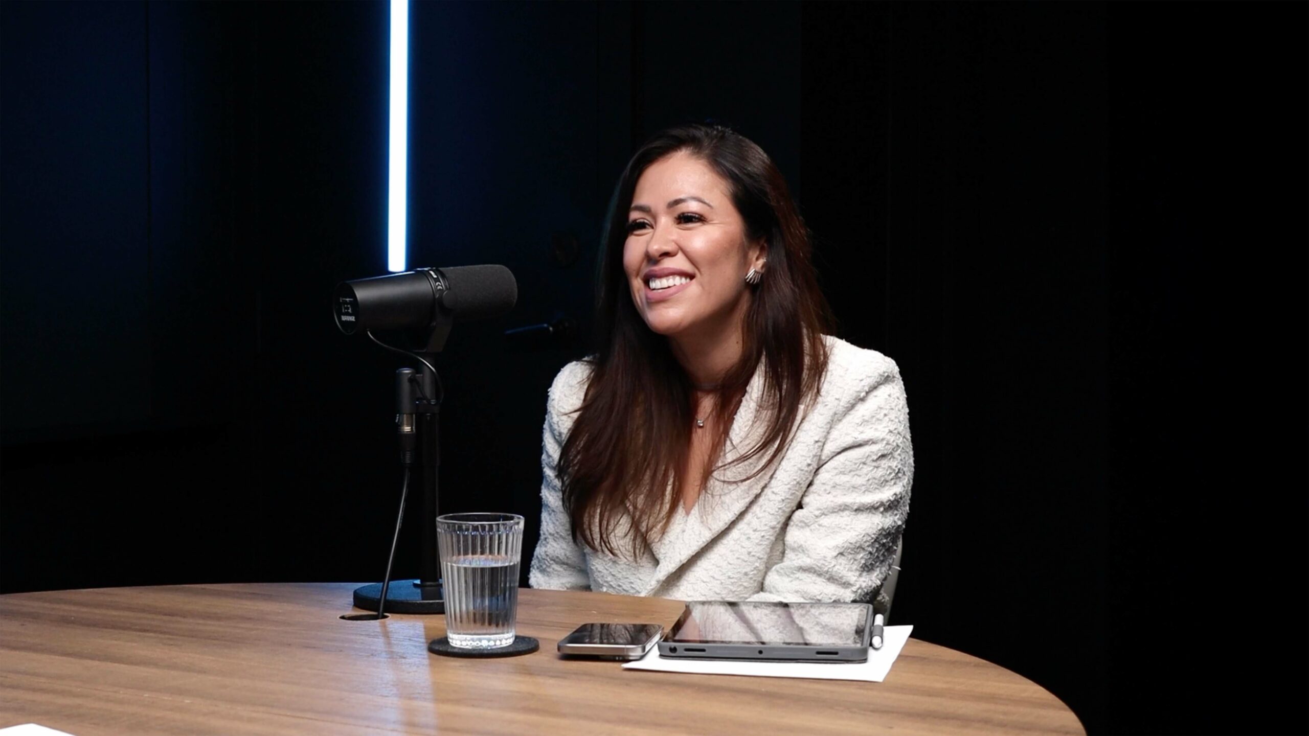 A woman in a white blazer sits at a round wooden table, speaking into a microphone, with a glass of water, a piece of paper, a phone and a tablet device nearby, against a dark background.