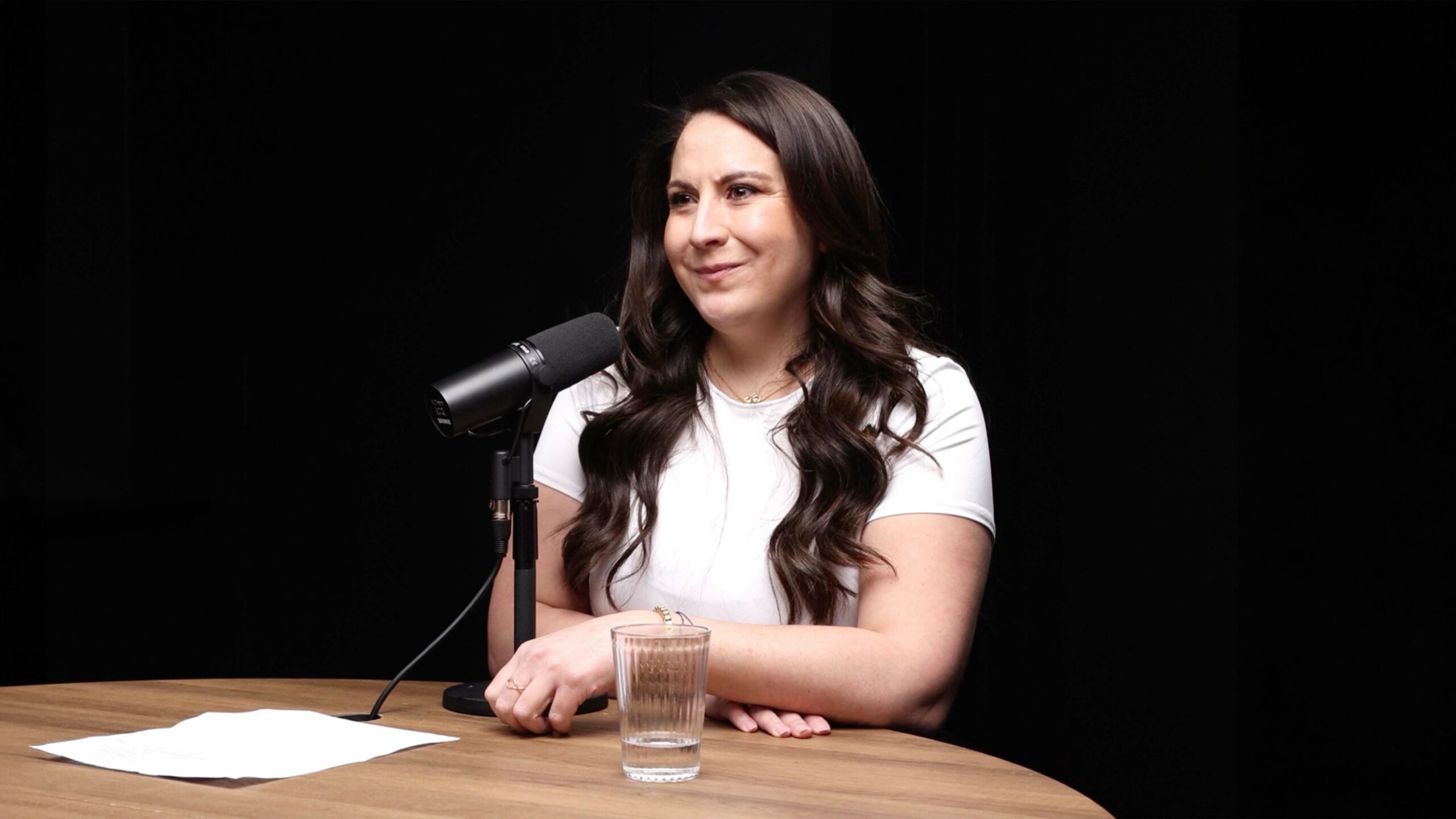 A person with long brown hair sits at a round wooden table, speaking into a microphone, with a glass of water and a piece of paper nearby, against a dark background.