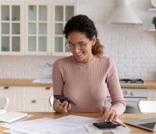 Young 30s Latin accountant woman work from home makes calculations on calculator, pay bills online via e-bank app on smartphone, manage household personal finances use modern tech. Bookkeeping concept